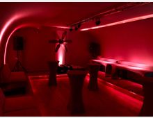 Ein kleiner, abgedunkelter Raum mit roten Lichteffekten, geschwungener Decke und Holzboden. Der Raum ist mit Stehtischen, einer Couch, Lautsprechern und einer zentralen Lüfterdekoration ausgestattet, die von einem Strahler beleuchtet wird.