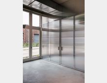 Innenraum mit glatten Betonboden, bodentiefen Kunststoff- oder Glastüren links und einer Wand aus transparenten Paneelen mit Metallrahmen rechts. Im Hintergrund sind durch die Fenster eine Backsteinfassade und ein Gehweg zu sehen.