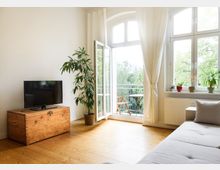 Wohnzimmer mit hellem Holzboden, einem grauen Sofa, einer Holztruhe mit Fernseher, und einem großen Fenster mit Glastür, die zu einem Balkon mit Blick auf grüne Bäume führt; Vorhänge sowie Zimmerpflanzen ergänzen die Einrichtung.