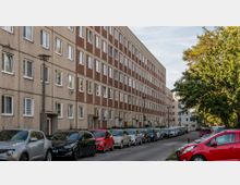 Wohnblock im Stil der Nachkriegsmoderne mit fünf Stockwerken und einer Fassade aus abwechselnd hellen und roten Paneelen; davor eine Straße mit parkenden Autos und rechts ein Baum mit grüner Krone.