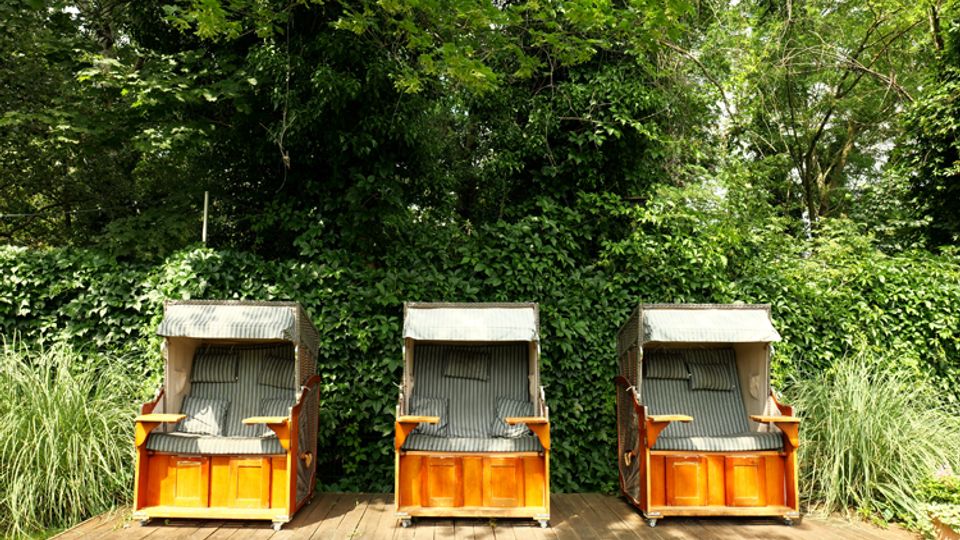 Drei Strandkörbe aus Holz mit gestreiften Polstern stehen auf einer Holzterrasse. Dahinter befinden sich dichtes grünes Laub, Sträucher und hohe Gräser, die den Bereich wie ein Garten oder Park umgeben.