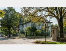 Eckhaus in einer ruhigen Wohngegend mit heller Fassade, umgeben von Gartenbepflanzung und Bäumen. Vor dem Haus stehen ein historischer Zaun, eine Straßenlaterne und Straßenschilder mit den Bezeichnungen „Willdenowstraße“ und „Malvenstraße“.