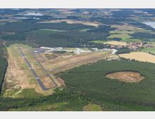 Luftaufnahme eines ländlichen Flugplatzes mit zwei asphaltierten Landebahnen, umgeben von Wald und Feldern. Im Hintergrund sind mehrere Gebäude des Flughafens sowie ein kleines Dorf und Seen in einer hügeligen Landschaft zu sehen.