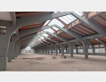 Innenraum einer großen, leerstehenden Industriehalle mit grauen Betonstützen und einem Dach aus Holz und Glas. Tageslicht fällt durch die großen Oberlichter ein, während die Halle einen offenen und ungenutzten Eindruck vermittelt.