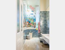 Kleines Badezimmer mit hellen Fliesenboden und türkisfarbenen Wandfliesen. Eine großflächige Wandgestaltung zeigt ein Unterwasser-Motiv mit bunten Meeresbewohnern; daneben befinden sich ein Fenster, ein Waschbecken, eine Toilette und Kinder-Badeaccessoires.