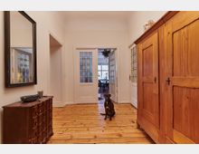 Heller Flur mit Holzboden, einem großen Holzschrank rechts und einer Kommode mit Spiegel links; im Hintergrund führen weiße Glastüren in einen weiteren Raum, in dem ein Esstisch sichtbar ist. Ein Hund sitzt auf dem Boden in der Mitte des Flurs.