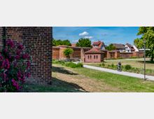 Grünanlage mit gepflastertem Weg vor einer langen, historischen Backsteinmauer und einem kleinen Backsteingebäude mit Walmdach. Im Hintergrund stehen Fachwerkhäuser und Bäume; links ist eine Backsteinwand mit blühenden Sträuchern zu sehen.