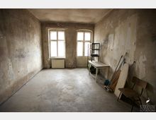 Ein leerer, abgenutzter Raum mit unverputzten Wänden, einem schmutzigen Betonboden und zwei großen Fenstern mit Holzrahmen. Im Raum stehen ein einfaches Regal, ein Tisch, eine Holzstuhllehne und mehrere zusammengelehnte Gegenstände wie ein Bügelbrett.