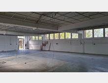 Leere Industrie- oder Werkstatthalle mit freiliegendem Raster der abgehängten Decke und herabhängenden Kabeln. Entlang der Wand befinden sich lange Fensterbänder mit Blick ins Grüne, ein offener Ausgang sowie weiße Wände und ein grauer Betonboden.