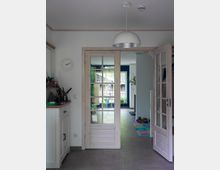 Blick in einen hellen Wohnraum mit halb geöffneter, verglaster Doppeltür aus Holz. Im Vordergrund steht ein weißer Schrank mit Dekorationsgegenständen, im Hintergrund sind ein farbiger Spielteppich und ein Raum mit blauer Wand zu sehen.