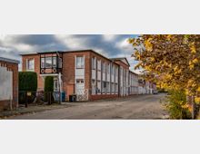Straßenszene mit einem langen Backsteingebäude mit weißen Fenstereinfassungen und einem kleinen hölzernen Erker. Auf der rechten Seite sind Herbstbäume mit gelben Blättern, und die Straße ist mit Laub bedeckt.