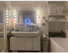 Gut beleuchtetes Badezimmer mit einem großen Waschbecken auf einem weißen Unterschrank, flankiert von Spiegeln mit Lampen. Handtücher sind ordentlich auf offenen Regalen gestapelt, und eine Badewanne ist rechts sichtbar. Ein runder, leuchtender Spiegel sowie dekorative Elemente ergänzen die moderne Einrichtung.