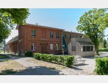 Backsteingebäude mit der Aufschrift "Festsaal" und Holzverkleidung im Anbau, daneben ein kleiner Außenbereich mit Kiesboden, Holztischen und einem grünen Sonnenschirm, umgeben von Büschen und Bäumen.