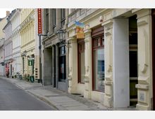 Straßenszene mit einer Reihe von historischen Gebäuden mit hellen Fassaden, verzierten Fenstern und Reklameschildern, darunter ein Weinladen und eine Bar. Kopfsteinpflaster und ein schmaler Gehweg säumen die Straße.