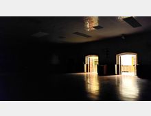 Ein dunkler, leerer Raum mit glänzendem Boden und einer abgehängten Decke mit rechteckigen Leuchten. Im Hintergrund sind zwei geöffnete Türen zu einem hell erleuchteten Bereich mit sichtbaren Wandelementen und Geländern.