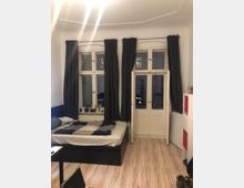 Ein Schlafzimmer mit einem Doppelbett, weißen Wänden und hellem Laminatboden. Drei hohe Fenster mit dunkelgrauen Vorhängen und eine Doppeltür mit Fenstereinsätzen dominieren den Raum; daneben ein weißes Regal mit roten und weißen Fächern.