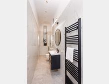 Schmales, modernes Badezimmer mit großformatigen marmorierten Wandfliesen und grauen Bodenfliesen, ausgestattet mit einer Glasdusche, wandhängendem WC und Waschtisch mit rundem Spiegel. Rechts ist ein schwarzer Handtuchheizkörper, die Decke hat eingelassene Spotlights und am Ende befindet sich ein schmales Fenster.