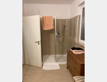 Ein schlichtes Badezimmer mit einer Eckdusche, die durch Glastüren abgetrennt ist. Neben der Dusche steht eine Holzkommode mit darauf gefalteten Handtüchern; eine weiße Tür und ein Fenster befinden sich ebenfalls im Raum.