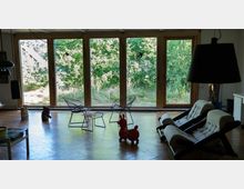 Wohnraum mit großen Holztüren, die zu einer Terrasse mit Blick auf einen grünen Garten führen. Im Innenraum stehen moderne Metallstühle, weiße Polstersessel und ein rotes Spielzeugtier auf einem Holzboden.