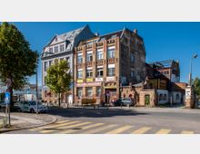 Straßenansicht mit einem dreistöckigen, historischen Backsteingebäude, das Ladenlokale wie „Propan-Discount“ beherbergt, daneben ein renoviertes Gebäude mit moderner Dachkonstruktion. Im Vordergrund eine Straße mit Zebrastreifen, geparkten Autos und Bäumen, rechts ein gemauerter Zaun mit Torbogen.