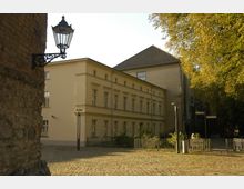 Gelbe Fassade eines klassischen, zweistöckigen Gebäudes mit großen rechteckigen Fenstern, flankiert von Bäumen auf der rechten Seite und einer mittelalterlichen Backsteinmauer mit einer Laterne auf der linken Seite. Der gepflasterte Platz im Vordergrund führt zu einem kleinen umzäunten Bereich mit einem Wegweiser und Hinweisschild.