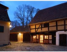 Ein gepflasterter Innenhof bei Dämmerung, umgeben von Fachwerkhäusern mit Holz- und Lehmwänden sowie Ziegeldächern. Im Hintergrund stehen unbelaubte Bäume vor einem blauen Abendhimmel, ein Gebäude ist von warmem Licht beleuchtet.