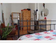 Interior of a bedroom with a black metal bedframe in the foreground, wooden wardrobe and open shelving against white walls, and a dresser with an octagonal wall mirror. The room has a hardwood floor with a patterned rug, a brown chair, a potted plant, and several coat racks and stored household items.