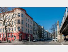 Straßenszene mit Wohngebäuden, darunter ein helles Eckhaus mit roten und beigefarbenen Fassaden und großen Fenstern. Rechts im Bild ist ein erhöhtes Eisenbahn-Viadukt zu sehen, flankiert von einer steinernen Säule mit dekorativen Elementen.