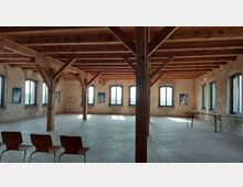 Großer Raum in einem historischen Gebäude mit sichtbaren Holzbalken an der Decke und unverputzten Backsteinwänden. Der Raum hat mehrere hohe Fenster mit Rundbögen, einige blaue Kunstwerke an den Wänden und einfache Holzstühle sowie Tische.