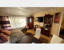 Wohnzimmer mit gemütlicher Einrichtung, darunter ein Tagesbett mit gemustertem Überwurf, ein Esstisch mit Stühlen, ein Holzschrank und ein Vitrinenschrank. Große Fenster mit transparenten Vorhängen lassen Tageslicht herein, und an der Wand hängt ein Flachbildfernseher sowie Dekorationsgegenstände.