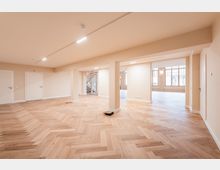 Großer, leerer Innenraum mit hellem Fischgrätparkett, beigen Wänden und mehreren Stützsäulen sowie Decken-Spotbeleuchtung. Im Hintergrund sind eine offene Treppe und große Sprossenfenster zu sehen, die viel Tageslicht in den Raum lassen.