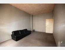 Ein schlichter, leerer Raum mit grauen Wänden und einem beigefarbenen Teppichboden. Im Vordergrund steht ein schwarzes Ledersofa, rechts im Bild befindet sich eine weiße Holztür.