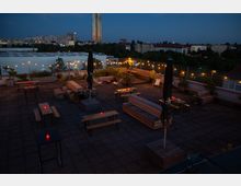 Beleuchtete Dachterrasse bei Dämmerung mit Holzbänken, Tischen und Pflanzen in Hochbeeten. Kleine rote Kerzen auf den Tischen sowie Lichterketten sorgen für stimmungsvolle Beleuchtung; im Hintergrund ist eine städtische Skyline zu sehen.