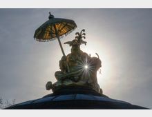 Eine goldene Statue, die eine sitzende Figur mit einem verzierten Kopfschmuck und einem Schirm über sich darstellt; sie befindet sich auf einem Dach mit einer dunkelblauen Kuppel. Die Sonne scheint hinter der Statue, wodurch sie als Silhouette hervorgehoben wird, während ein Lichtstrahl durch ihre Oberfläche reflektiert wird.