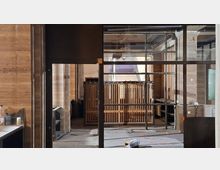 Innenraum eines im Umbau befindlichen Gebäudes mit Holzpaneelwänden und großen, schwarz gerahmten Glas- und Metalltrennwänden. Im Hintergrund stehen Holzgeländer und offene Deckeninstallationen, am Boden liegen Bau- und Verpackungsreste.