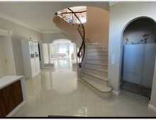 Innenansicht eines hellen Wohnbereichs mit einer geschwungenen Treppe, deren Geländer aus dekorativem Metall besteht. Der Raum hat glänzende Fliesenböden, eine Tür mit Durchgangsbogen, helle Wände und große Fenster, die viel Tageslicht hereinlassen.