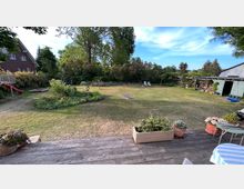 Großer Garten mit Rasenfläche, einem runden Blumenbeet in der Mitte und hohen Bäumen im Hintergrund. Rechts steht ein kleines Gartenhaus, links ein rotes Spielgerät, und im Vordergrund ist eine Holzterrasse mit Blumenkästen zu sehen.