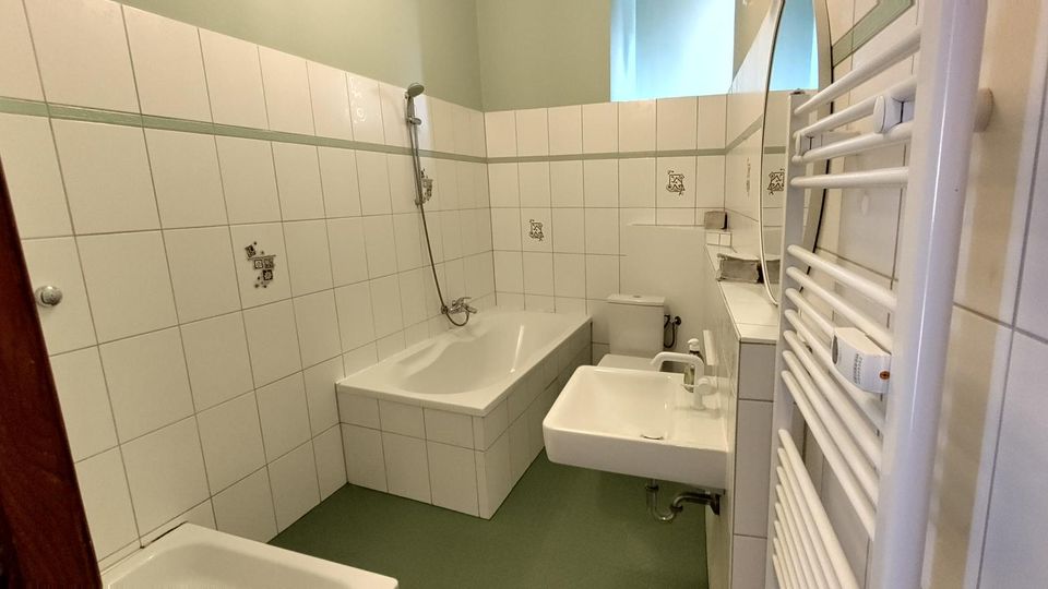 Kleines, gefliestes Badezimmer mit Badewanne und Duscharmatur an der Rückwand, WC und wandhängendem Waschbecken. Rechts befindet sich ein großer Handtuchheizkörper, darüber ein Fenster für Tageslicht.