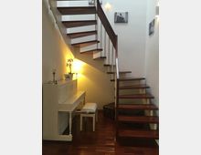 Eine moderne Holztreppe mit offenen Stufen und Metallgeländer führt nach oben, flankiert von einer weißen Wand. Darunter stehen ein weißes Klavier mit passendem Hocker, eine Stehlampe sowie ein Koffer auf einem Holzboden, und an den Wänden hängen einige Schwarz-Weiß-Fotografien.