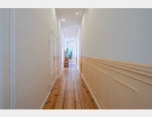 Langer, schmaler Flur mit Holzboden, weißen Wänden und heller Wandvertäfelung. An den Seiten befinden sich Türen, und am Ende des Flurs stehen ein Holzstuhl sowie Pflanzen in einem beleuchteten Bereich.