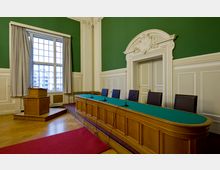 Ein Sitzungssaal mit grünen Wänden, hellen Zierleisten und einem großen Fenster mit grauen Vorhängen. Im Vordergrund befindet sich ein langes hölzernes Podium mit grüner Oberfläche, ausgestattet mit Mikrofonen, und daneben ein erhöhtes Rednerpult.