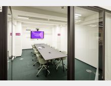 Besprechungsraum mit einem langen rechteckigen Tisch und mehreren hellgrünen Stühlen auf Rollen, umgeben von weißen Wänden und Glaswänden. Im Hintergrund hängt ein großer Bildschirm mit Text, der den Raum beschreibt.