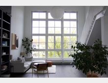 Wohnzimmer mit hohen Decken und großen Fensterfronten im Rasterdesign, die viel Tageslicht hereinlassen. Der Raum ist modern eingerichtet mit einer grauen Couch, Holzmöbeln und mehreren großen Zimmerpflanzen; eine weiße Treppe führt nach oben.