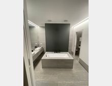 Modernes Badezimmer mit grauen Fliesen, einer freistehenden Badewanne in der Mitte und einer dunklen Wand als Akzent. Links befindet sich ein langer Waschtisch mit zwei weißen Waschbecken, und rechts führt eine Tür zu einem weiteren Bereich.