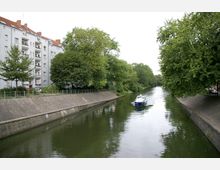 Ein schmaler Kanal mit grünen Ufern, flankiert von Bäumen auf beiden Seiten; links steht ein Mehrfamilienhaus mit Balkonen, während ein kleines Boot den Kanal entlangfährt.