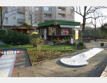Kleiner Minigolfplatz mit Betonbahnen und roten Begrenzungen, umgeben von bepflanzten Beeten und einer gepflasterten Fläche. Im Hintergrund steht ein Kiosk mit grünem Dach und einer gestreiften Markise, daneben Wohngebäude und Bäume.