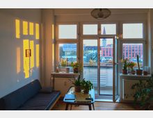 Wohnzimmer mit großen Fenstern, durch die warmes Sonnenlicht hereinfällt und auf die Wand Schatten wirft. Das Zimmer ist mit einer Couch, einem Tisch mit Zimmerpflanzen und Zugang zu einem Balkon ausgestattet, der den Blick auf eine städtische Umgebung mit Gebäuden und einem Kirchturm bietet.