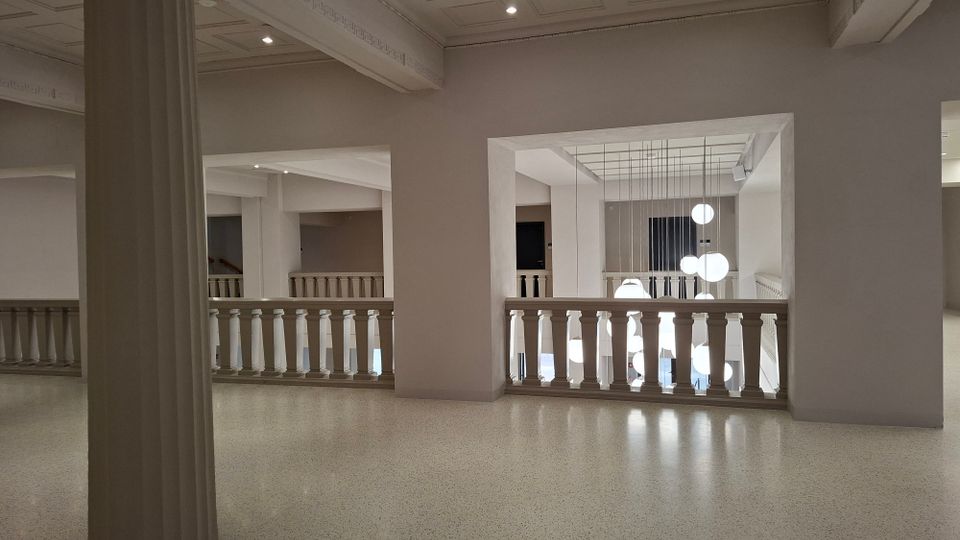 Innenraum eines modernen Museums oder Foyers mit weißen Wänden, Balustraden und einem offenen Atrium. In der Mitte hängen mehrere kugelförmige Pendelleuchten, während Säulen und breite Deckenbalken die Architektur gliedern.