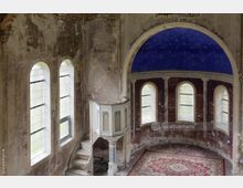 Verlassener Kirchenraum mit abblätternden Wänden, einer blauen, halbrunden Kuppeldecke sowie einer Kanzel mit einer schmalen Wendeltreppe. Die Fenster sind schmal und im Rundbogenstil, der Boden ist mit einem roten Teppich belegt.