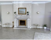 Ein eleganter Wohnraum mit einem dekorativen Kamin aus weißem Marmor, verziert mit feinen Schnitzereien. Der Kamin ist von hellen Wänden mit floraler Tapete umgeben, und darüber hängt ein rechteckiger Spiegel in einem goldenen Rahmen; daneben befinden sich Wandlampen sowie kleine Beistelltische und Pflanzen.